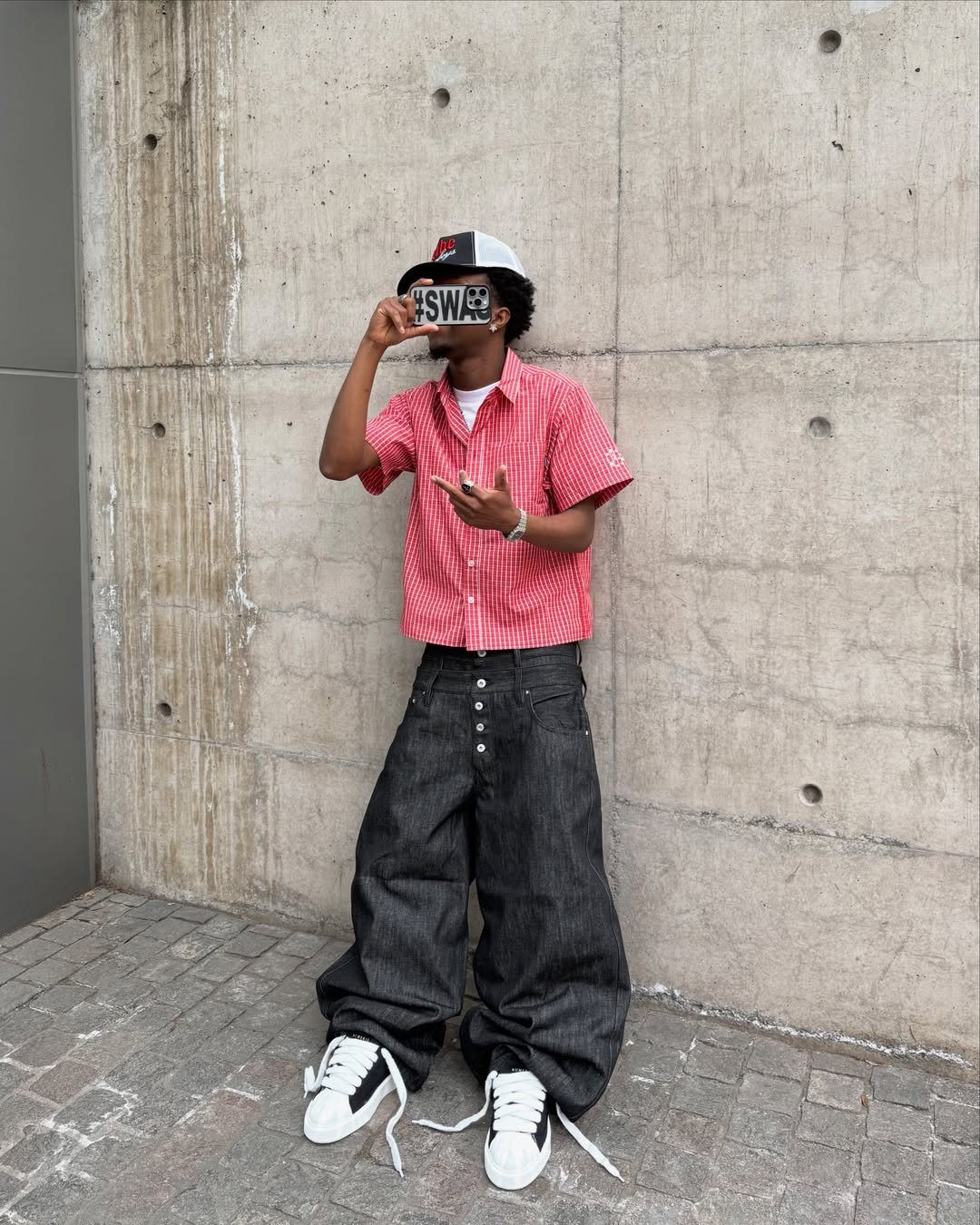 A person in baggy black pants and white chunky sneakers standing against a concrete wall.