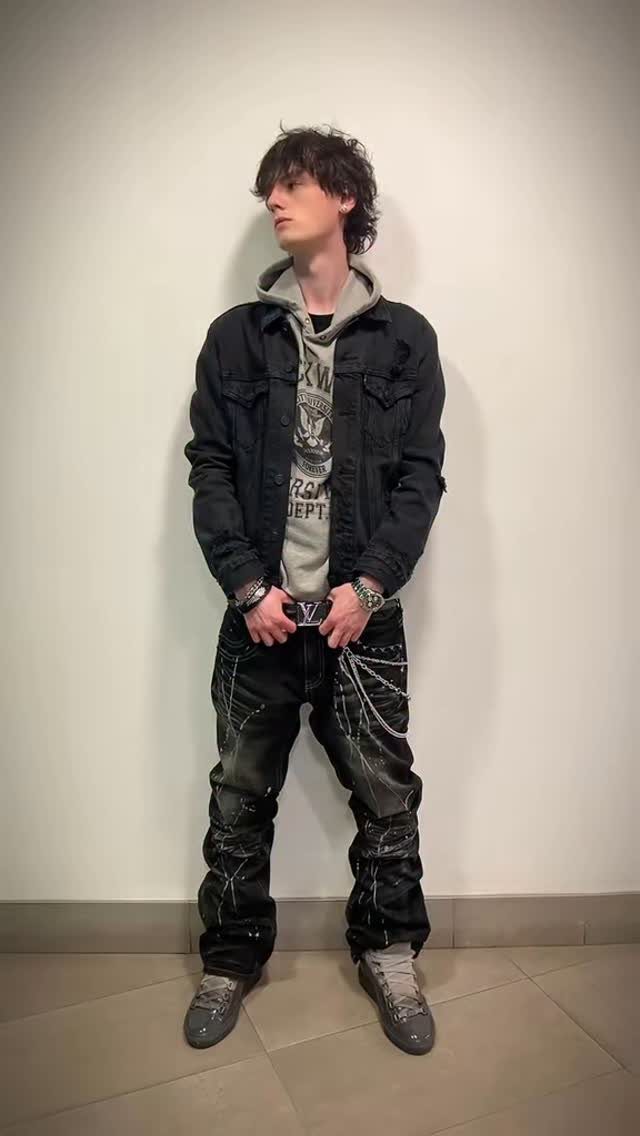A young man posing in a black jacket, graphic hoodie, and splattered black jeans.