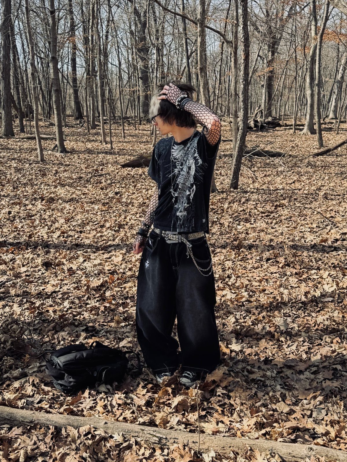 A person in pop-punk style clothing stands in a forest of bare trees and fallen leaves.