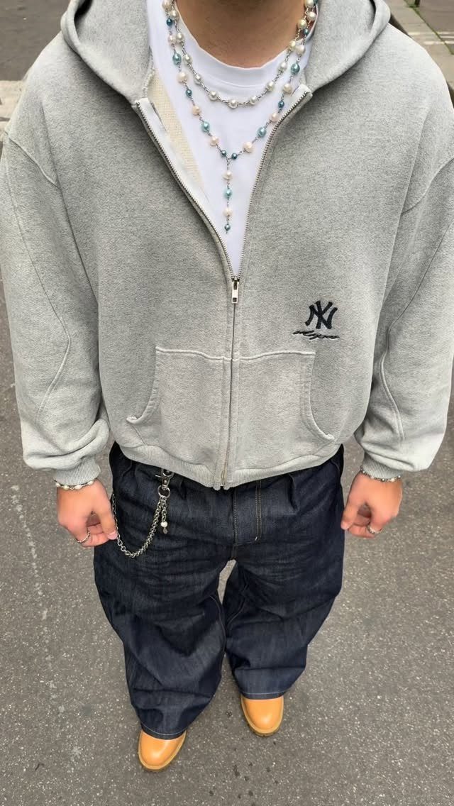 A person wearing a grey zip-up hoodie, baggy dark jeans, and layered pearl necklaces.