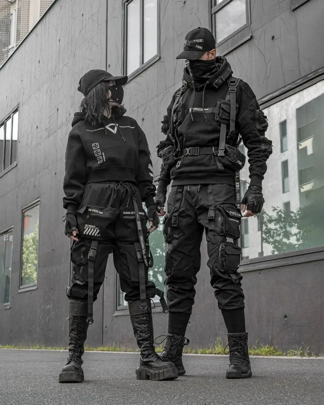 Two people posing in black tactical techwear outfits with straps and combat boots outdoors.