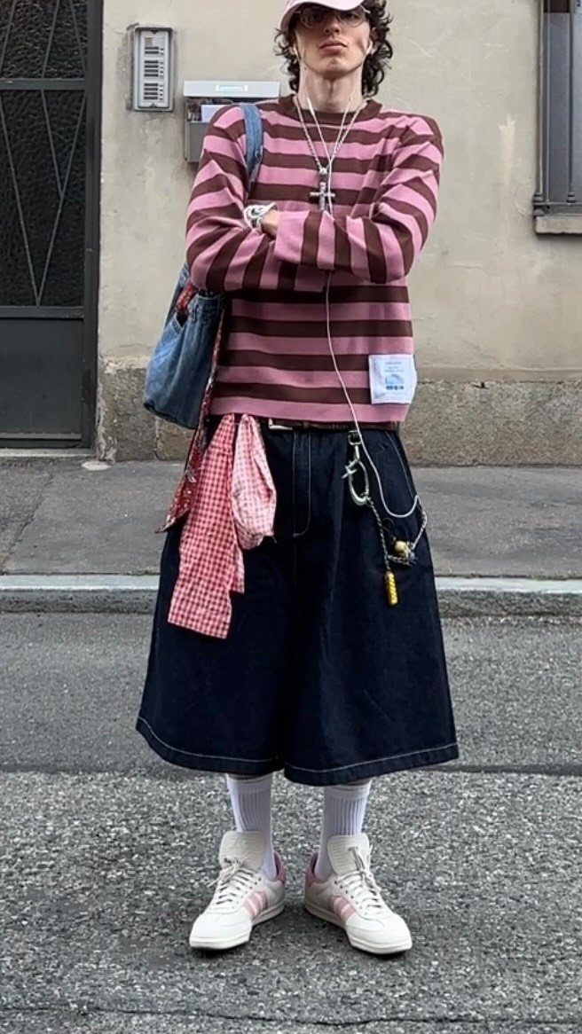 A person posing in a striped shirt, wide denim shorts, and pink sneakers with silver chains.