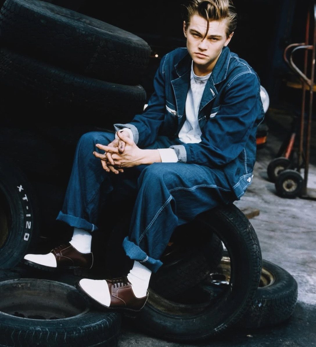 Young man in a blue denim jumpsuit sitting on tires in a dark garage setting.