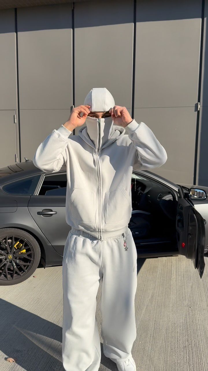 Person wearing a white hooded tracksuit and cap standing in front of a grey car.