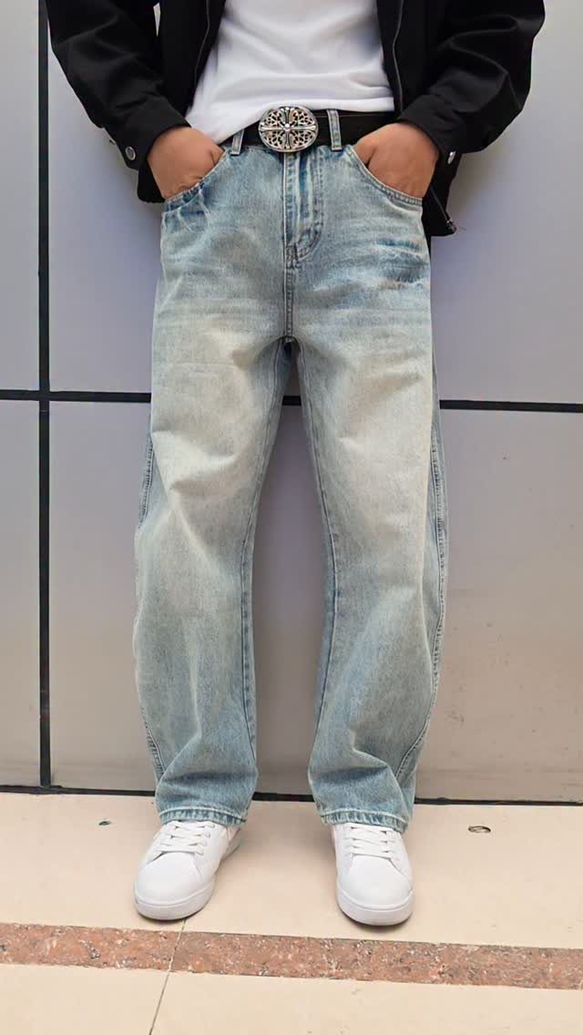 Person wearing baggy light blue denim jeans, a black jacket, and white sneakers against a wall.