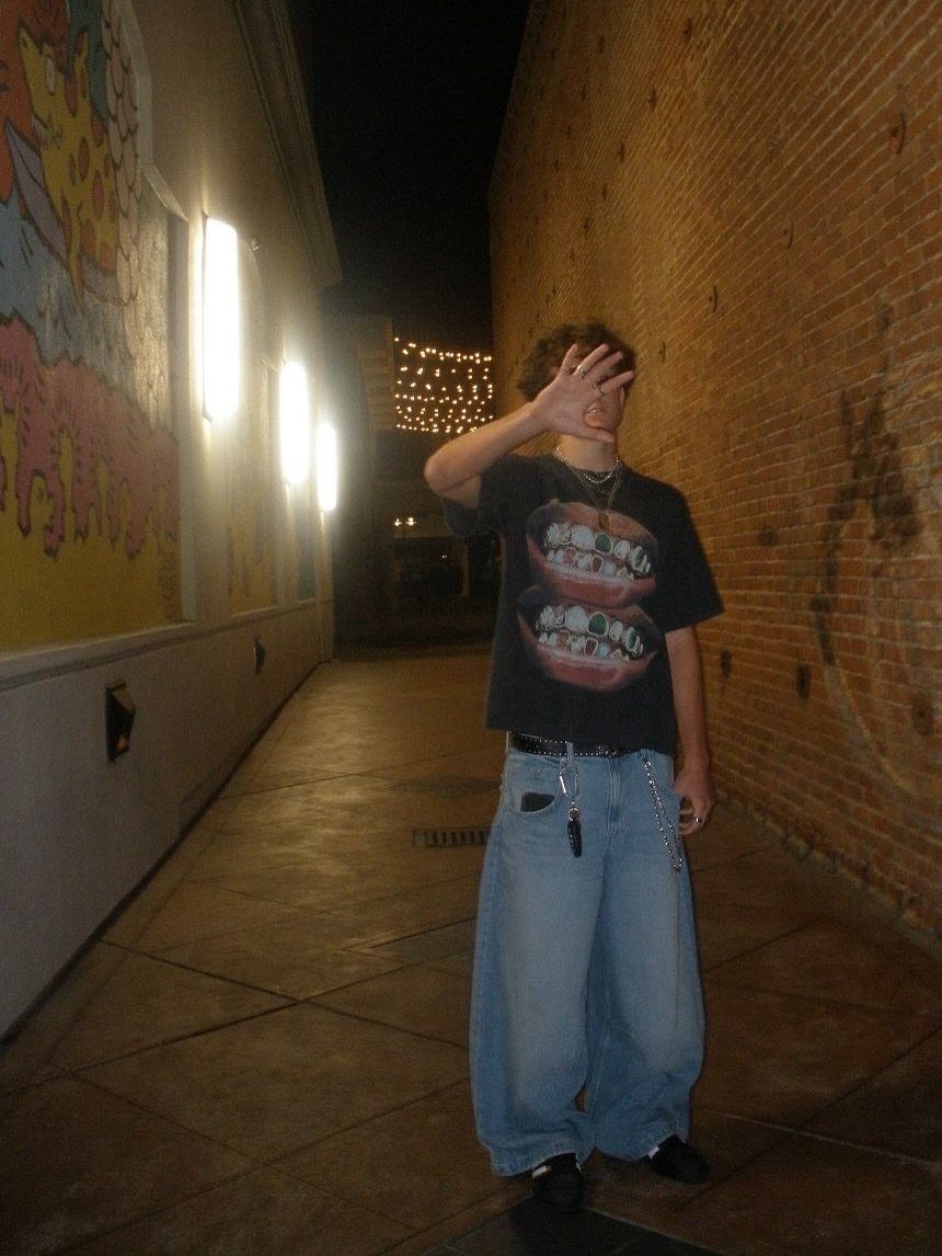 A person wearing a graphic t-shirt and baggy blue jeans standing in a dark alleyway.