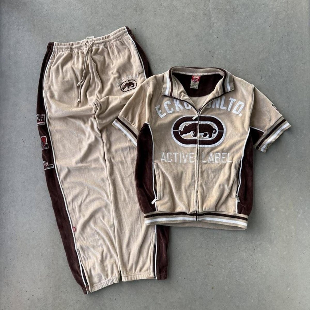 A beige and brown velour tracksuit set with branding laid flat on concrete.